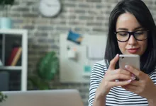 Una donna con gli occhiali guarda lo smartphone, seduta davanti a un portatile aperto