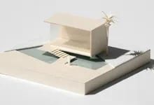 Modellino architettonico in cartoncino color panna: un padiglione dal tetto piano affacciato su una piccola piscina con scaletta, una palma di carta e una figura in miniatura, su una base quadrata