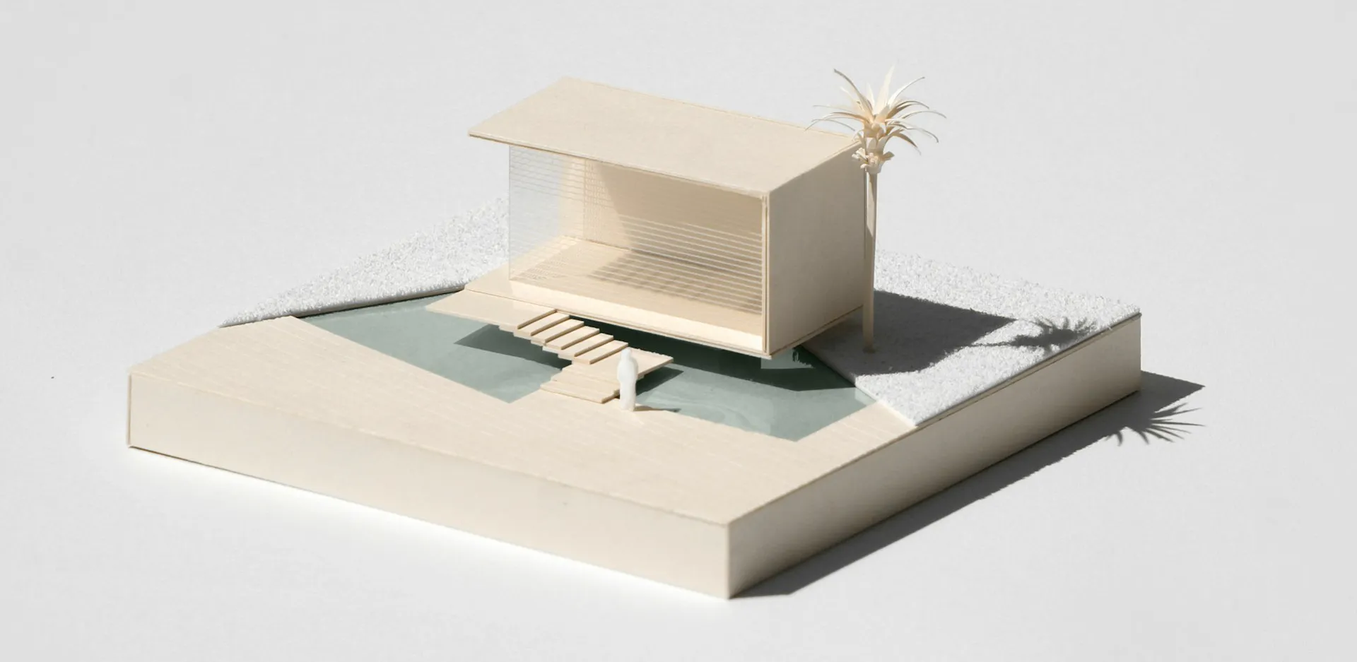 Modellino architettonico in cartoncino color panna: un padiglione dal tetto piano affacciato su una piccola piscina, con una palma di carta e una figura in miniatura su una base quadrata